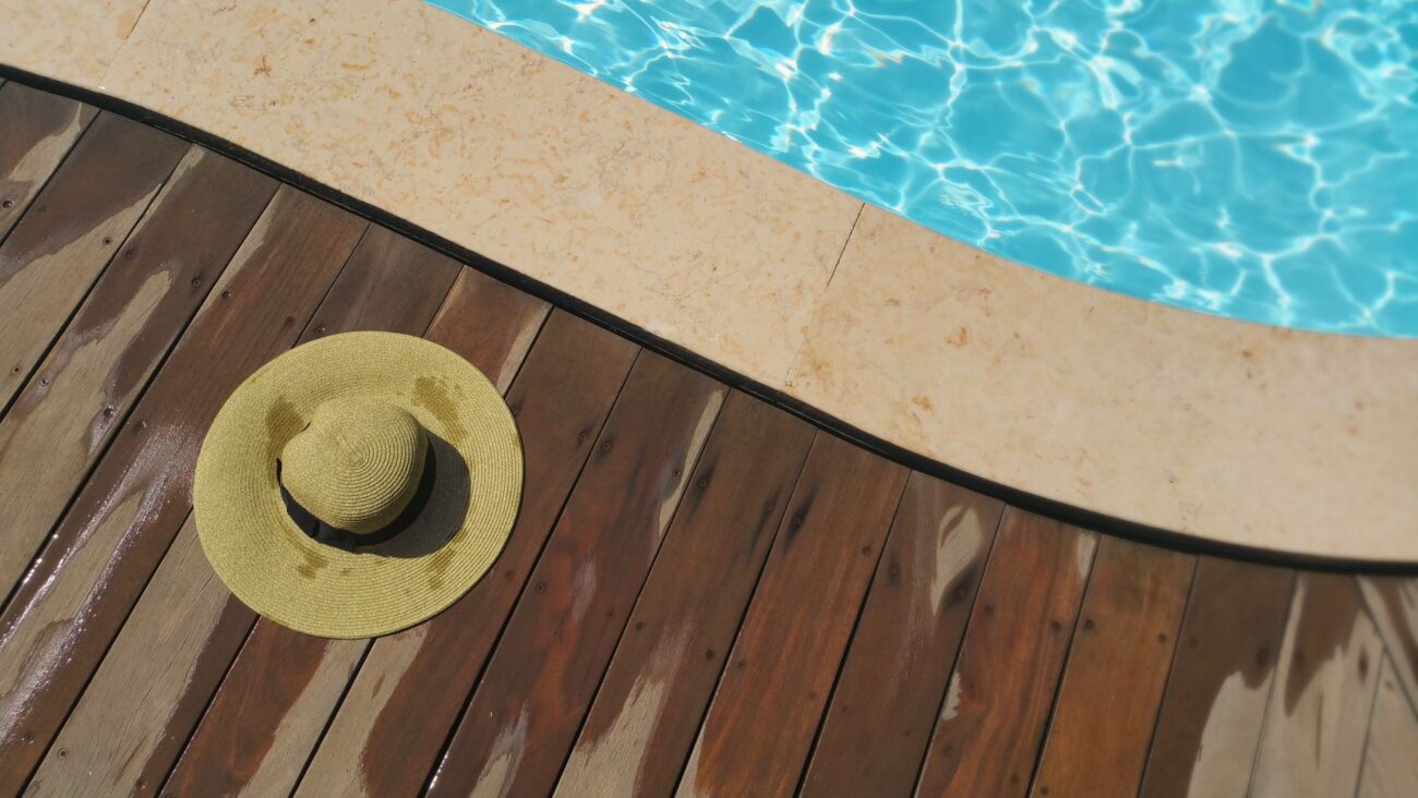 A straw hat rests on a wooden deck beside a sparkling blue swimming pool, perfect for summer relaxation. – deck building company 2026