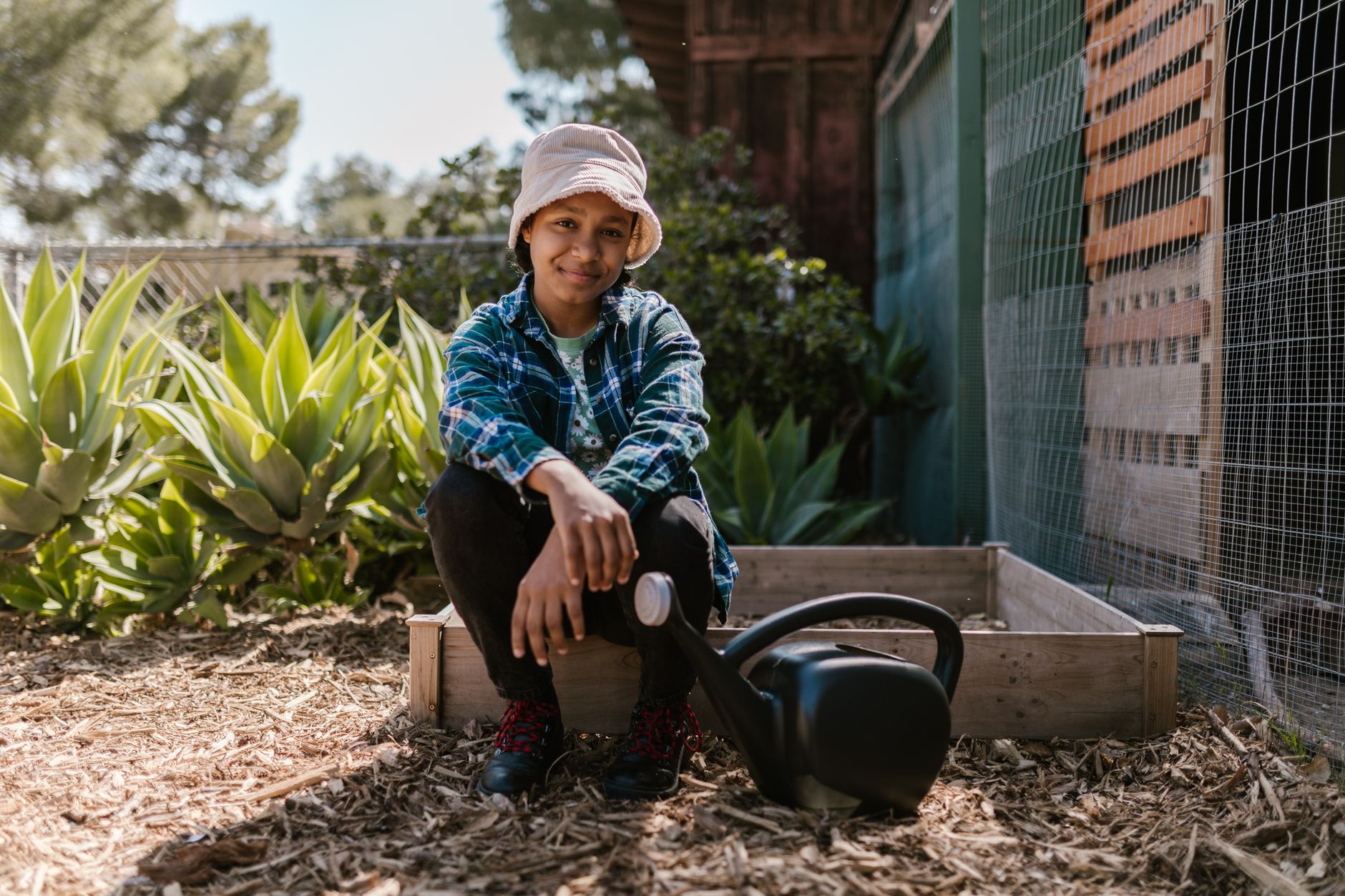 A young gardener with a watering can sitting in a sunlit outdoor garden, wearing a hat and plaid shirt.