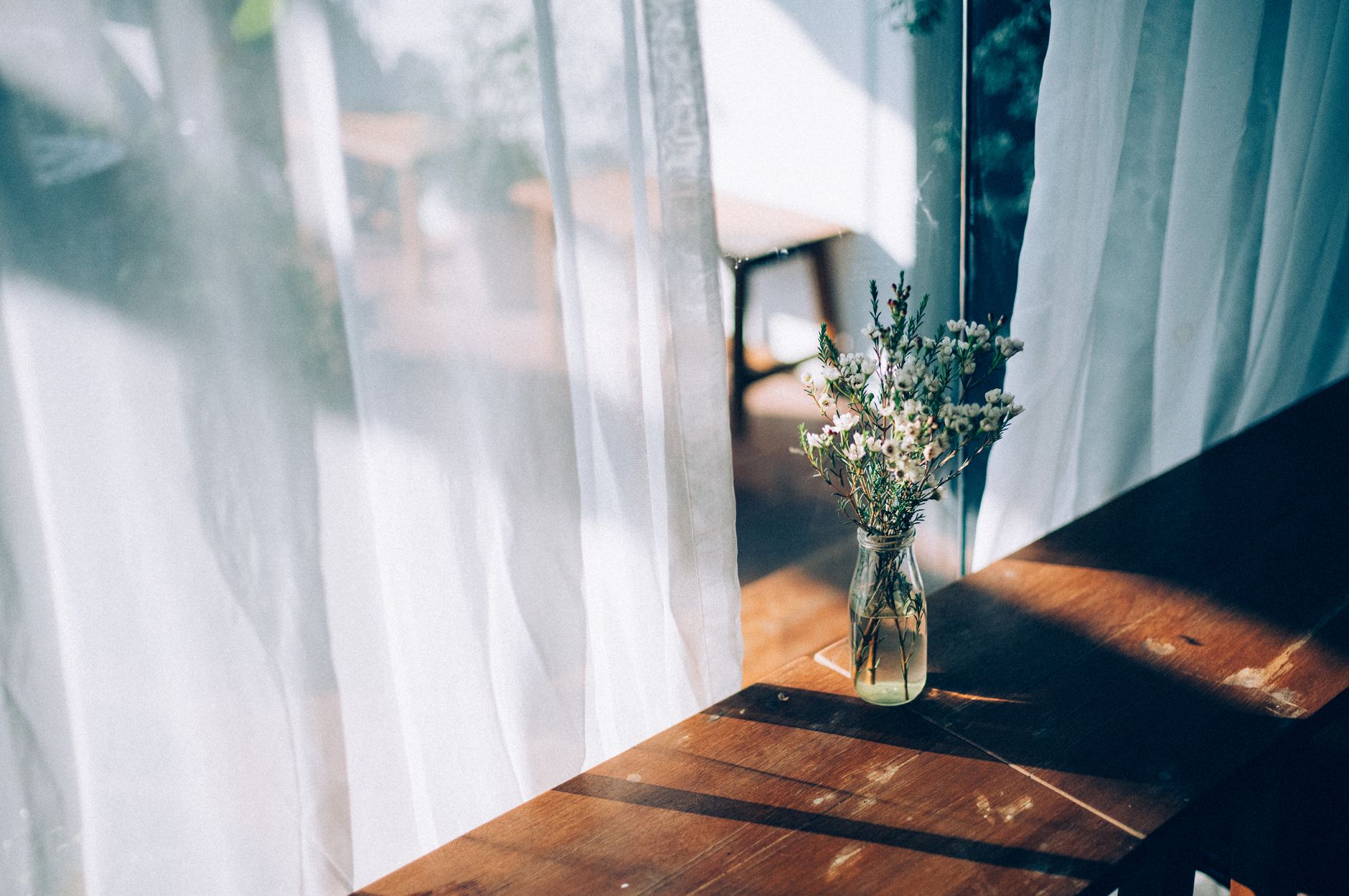 A rustic glass vase with flowers on a sunlit wooden windowsill against sheer curtains.