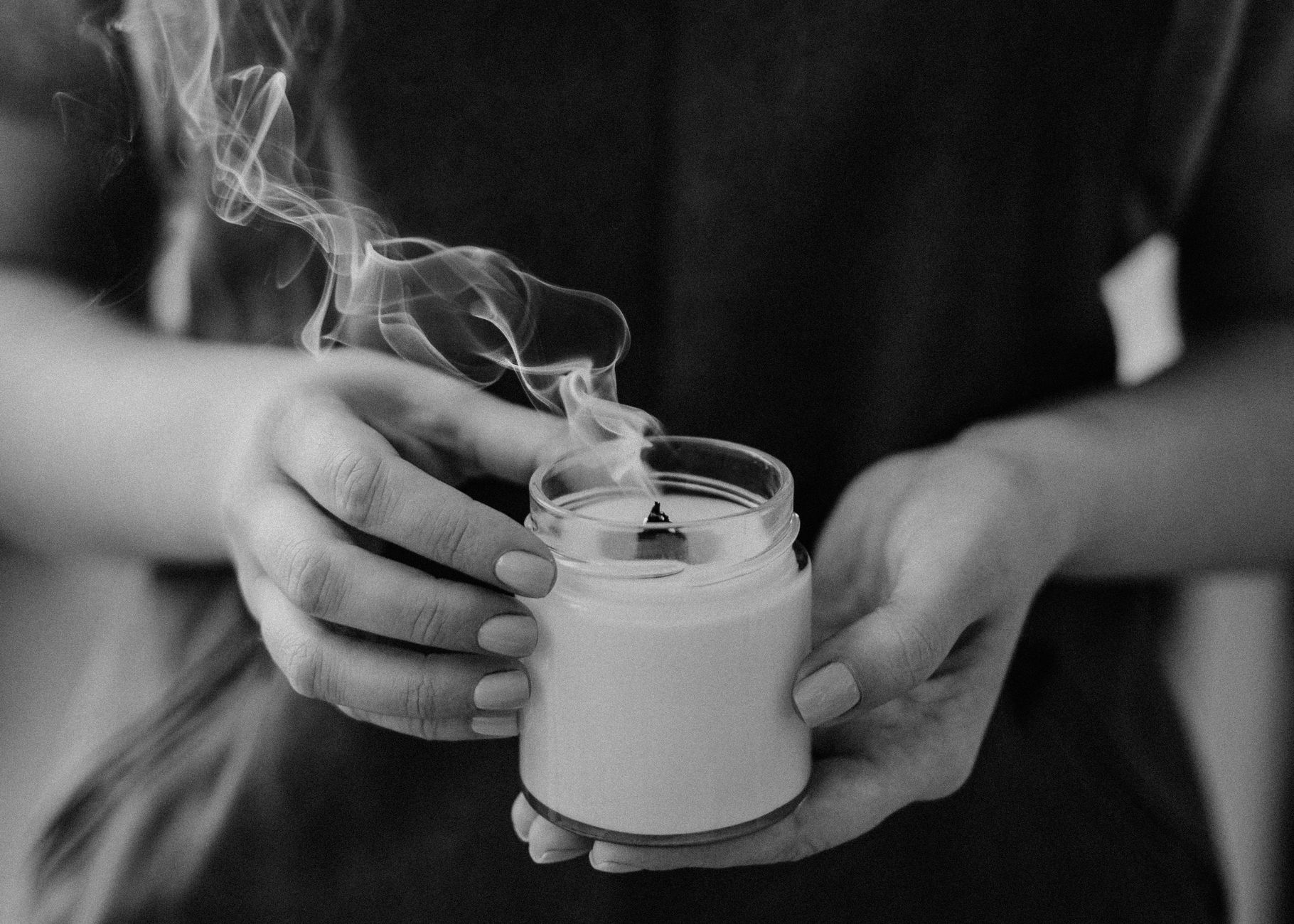 A close-up black and white photo of hands holding a jar candle with smoke.