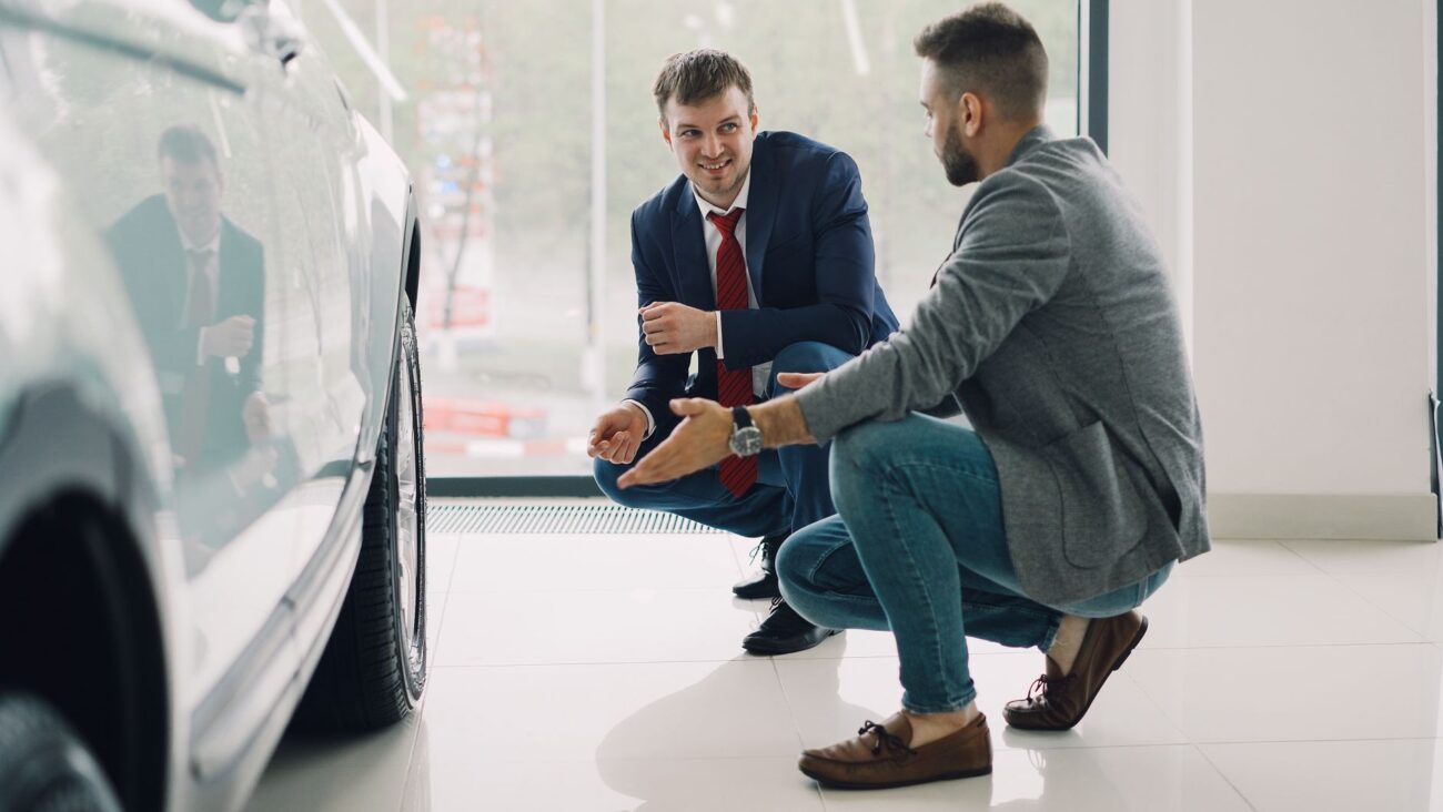 Two men discussing car features in a showroom, kneeling near a vehicle. – car dealers probably don
